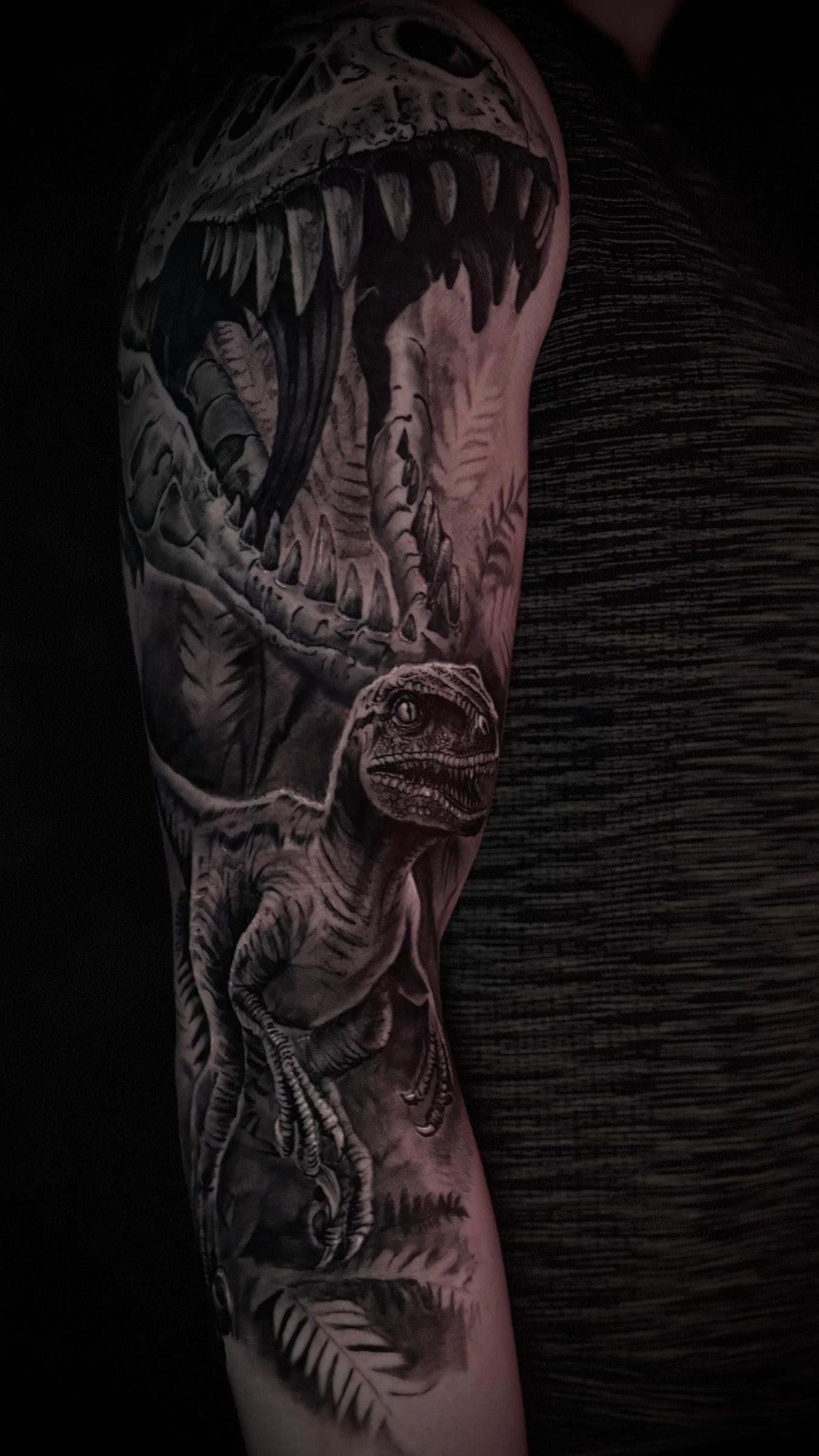 Black and grey tattoo of a dinosaur skull with open jaws above a raptor among ferns, running down an arm.