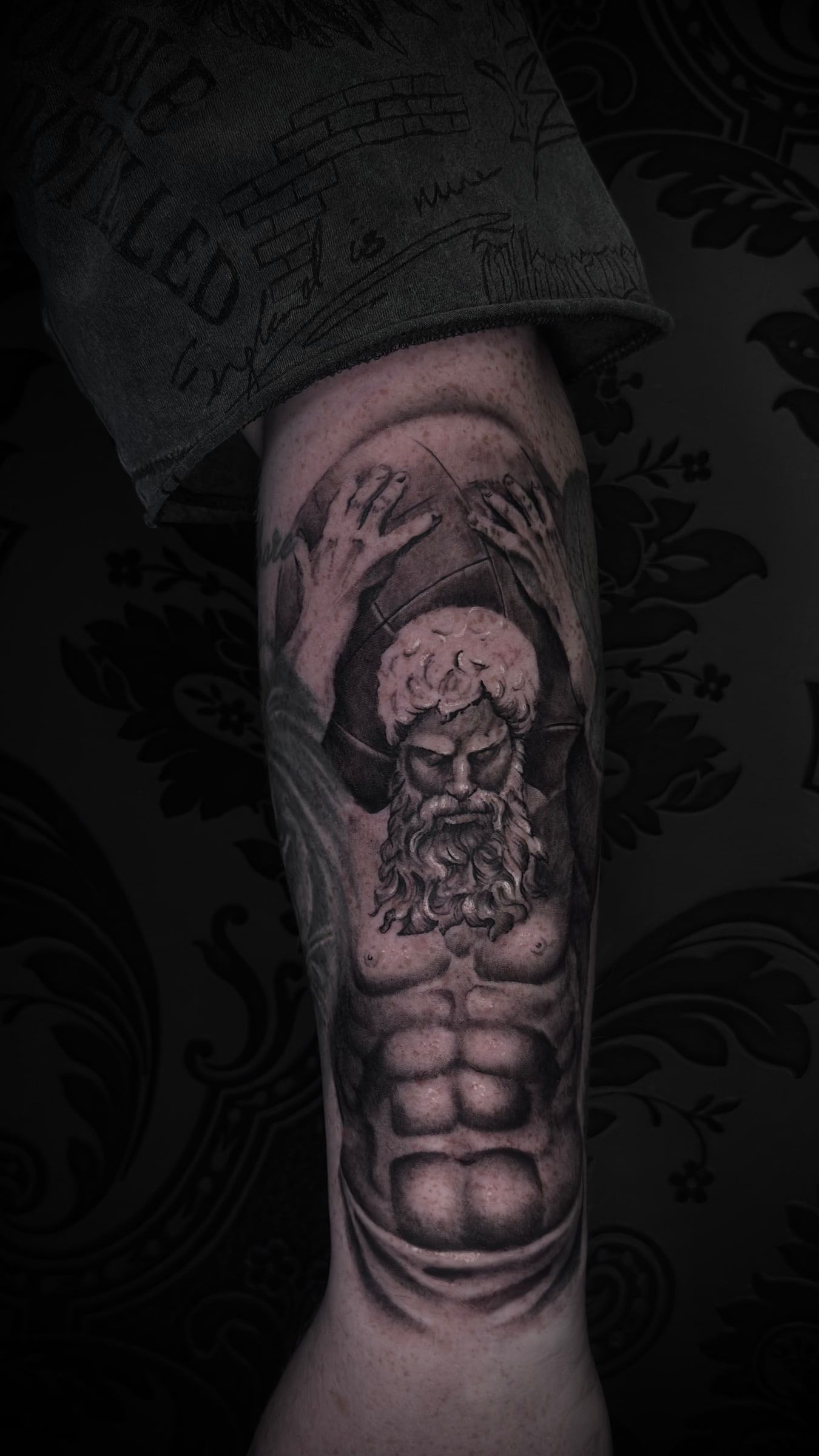 Black and grey tattoo of a bearded figure with both arms raised, bearing a weight overhead, on an upper arm.