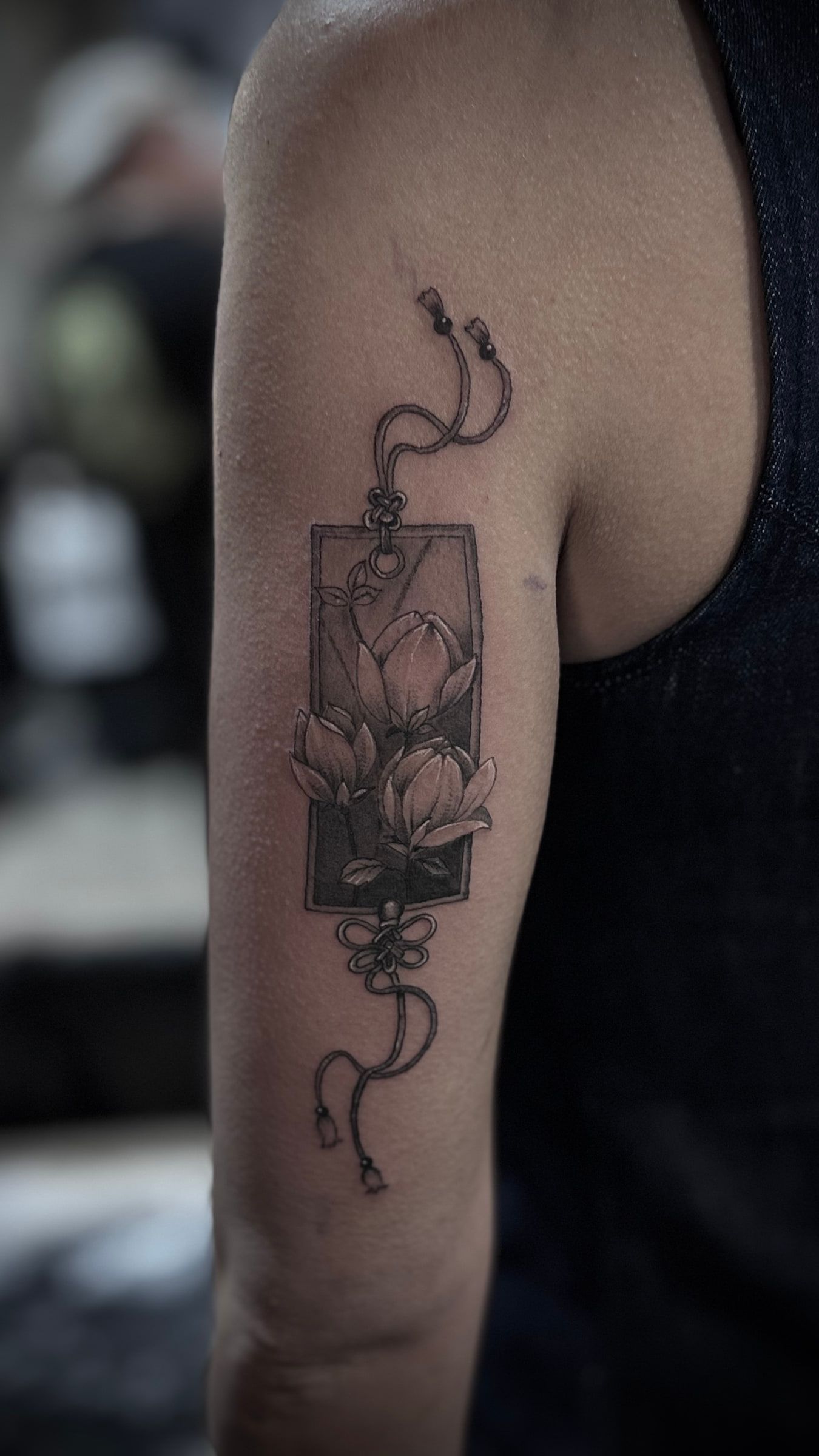 Fine line black and grey tattoo of flowers in a rectangular frame hung from a knotted cord, on an upper arm.