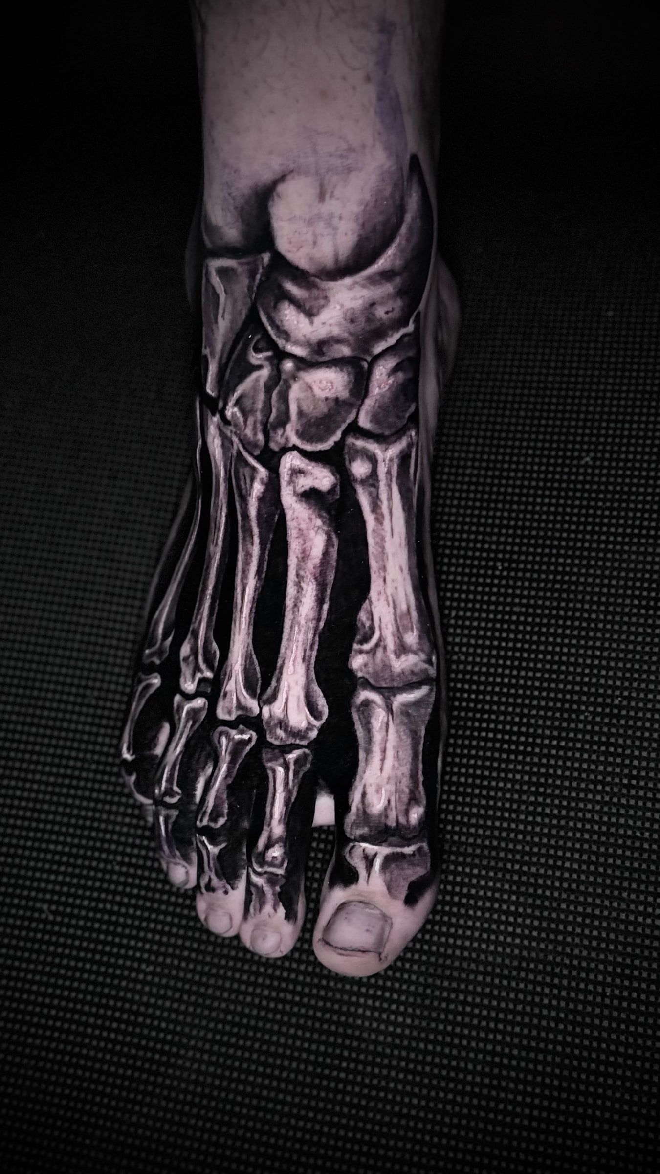 Black and grey tattoo of the bones of the foot, picked out against solid black, across the top of a foot.