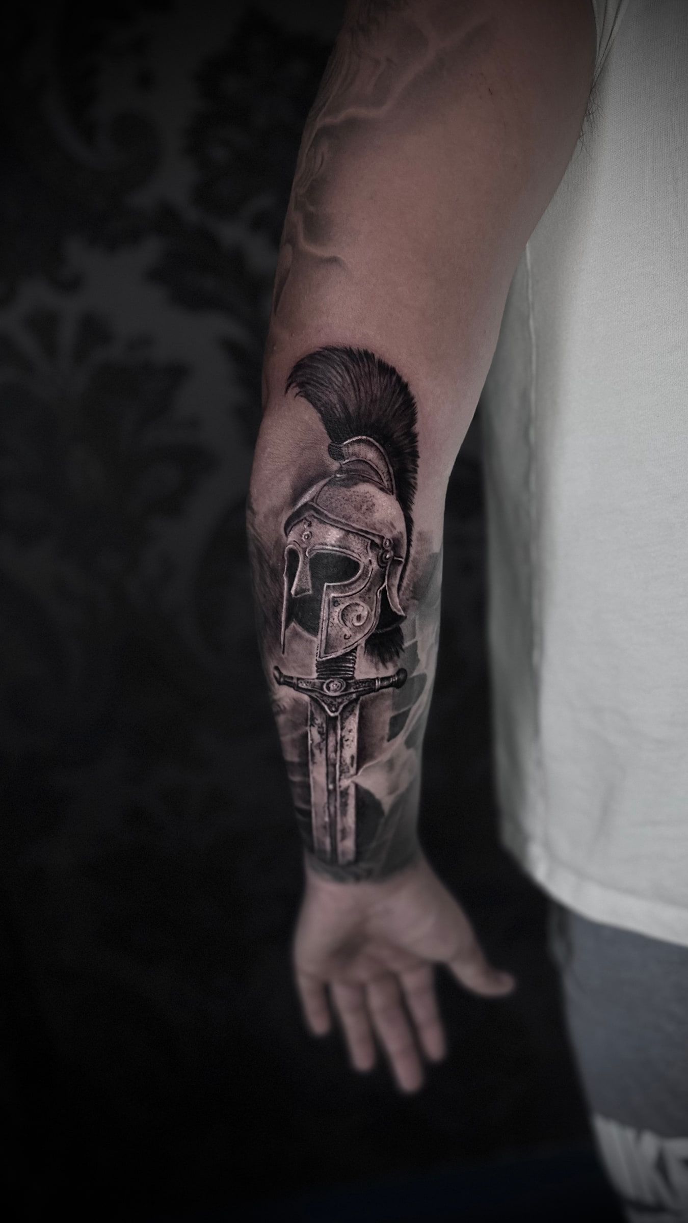 Black and grey realism tattoo of a crested helmet above a sword, on a forearm.
