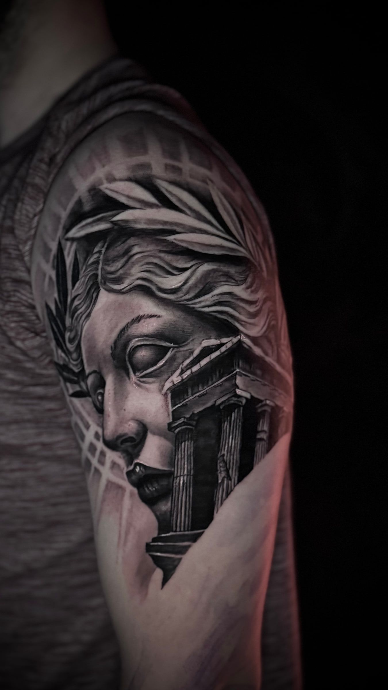 Black and grey tattoo of a laurel-wreathed classical statue with a ruined colonnade across the lower half of the face, on an upper arm.