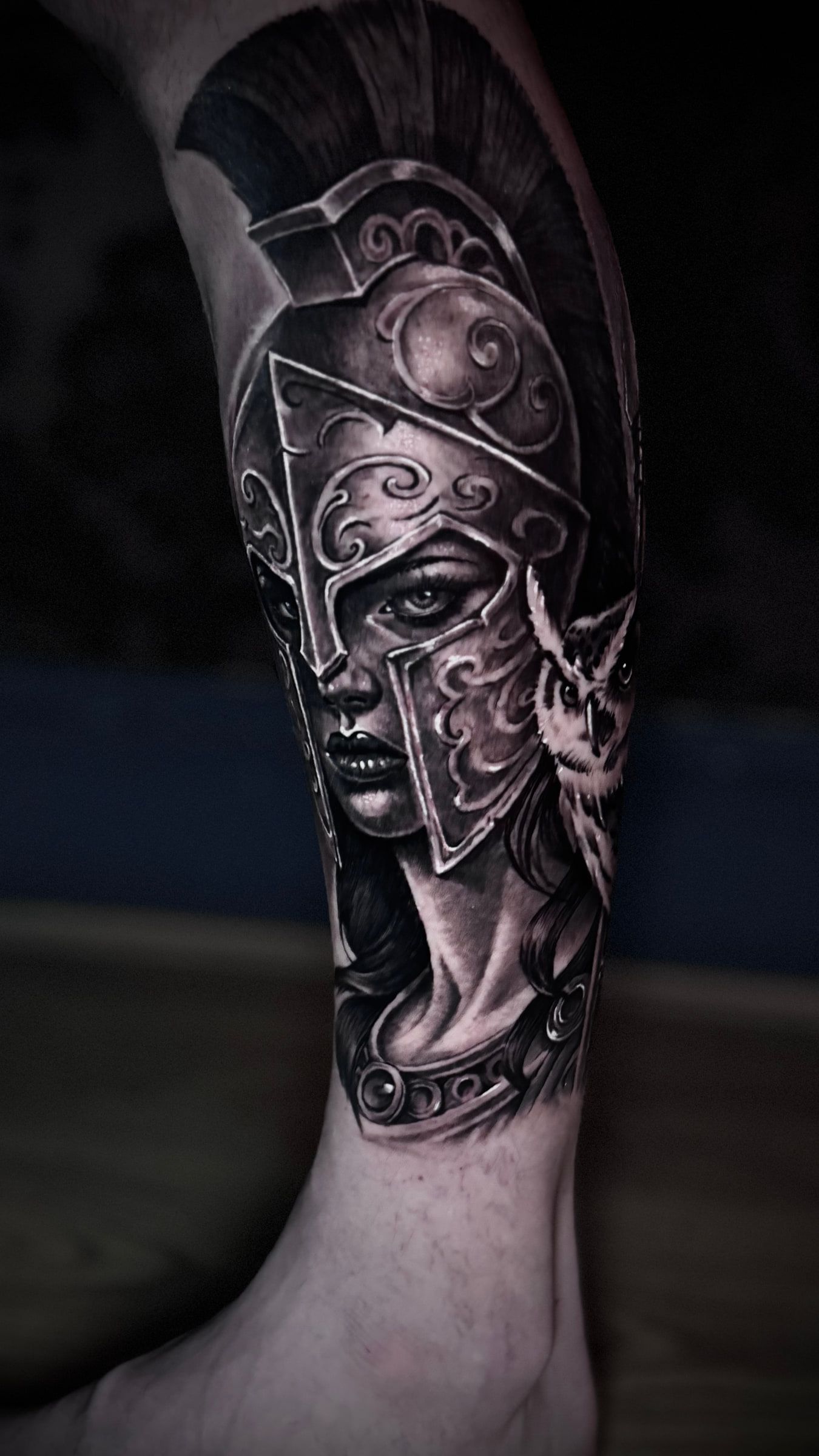 Black and grey realism tattoo of a helmeted female warrior with an owl at her shoulder, on a calf.
