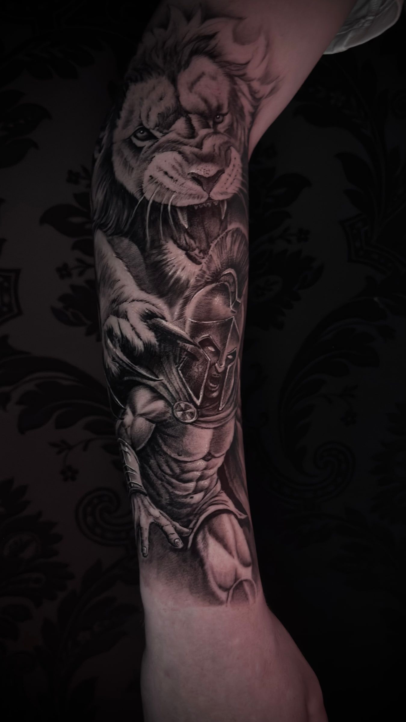 Black and grey realism tattoo of a roaring lion above a helmeted warrior in armour, on a forearm.