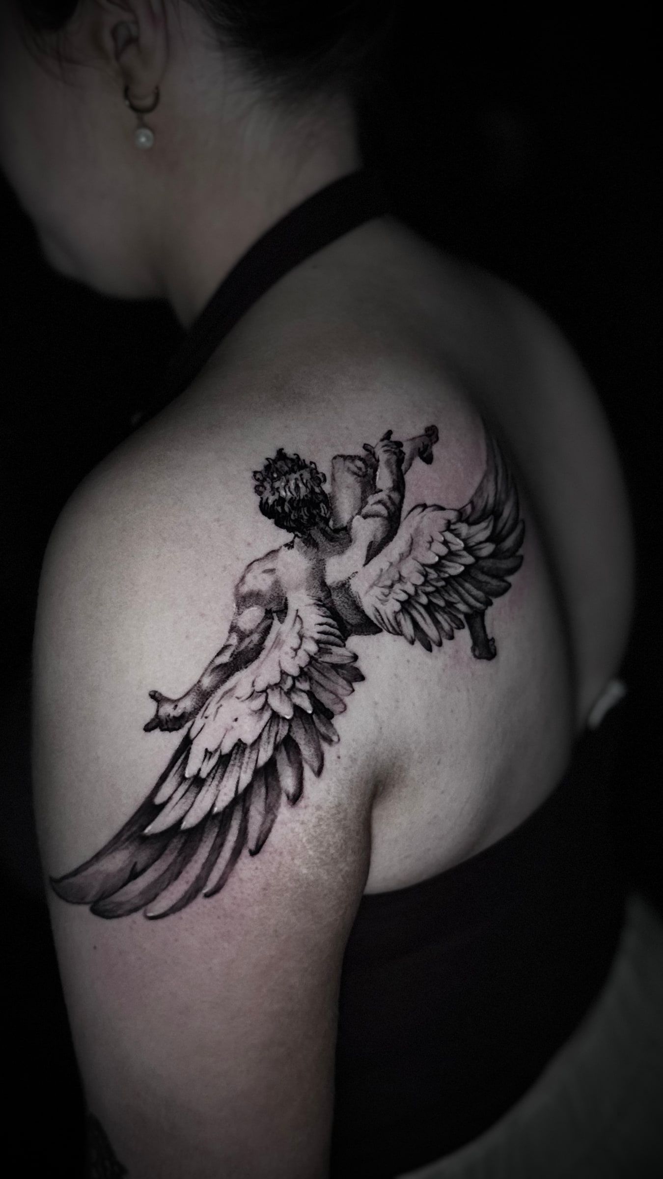 Black and grey tattoo of a winged cherub raising a horn, across a shoulder.